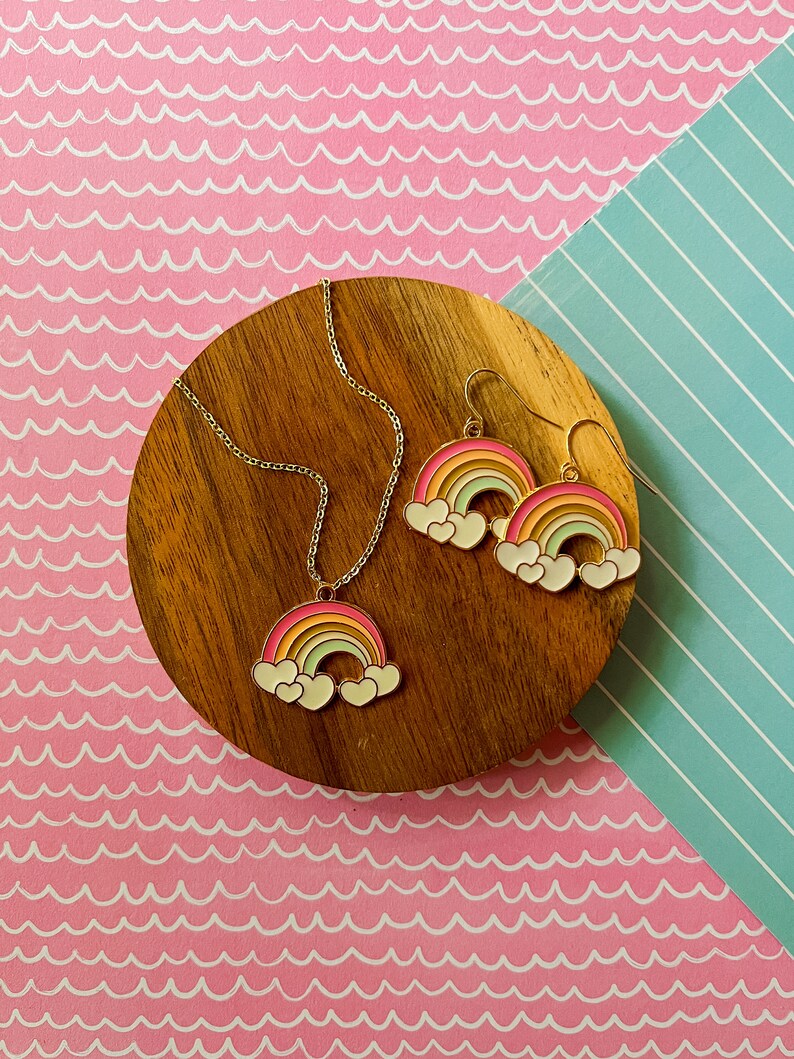 May include: A gold necklace and earrings set featuring rainbow and cloud charms. The necklace has a delicate chain and the earrings are dangle style.