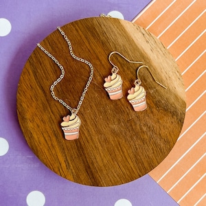 May include: A gold necklace and earrings set with cupcake charms. The cupcake charms are pink, white, and yellow with a pink heart on top.