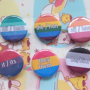 May include: A collection of nine circular pin-back buttons featuring various pride flag colour patterns and gender pronouns including she/her, he/him, they/them, it/its, hij/hem, die/hun, and zij/haar, along with a yellow button with a purple circle.