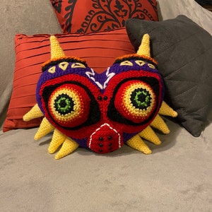 Puede incluir: Una almohada de crochet con forma de la Máscara de Majora de la serie de videojuegos The Legend of Zelda. La máscara es roja, amarilla, morada y verde con ojos negros y una boca blanca.