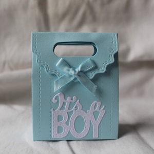 Pode incluir: Uma sacola de presente azul claro com um laço branco e o texto "It's a Boy" em branco.