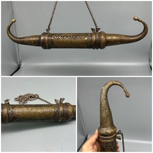 May include: An elongated, cylindrical brass pen case featuring intricate filigree cutouts and detailed Arabic calligraphy engravings. The object has curved, pointed ends and is suspended by a metal chain attached to two small loops.