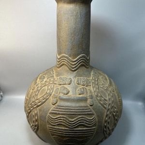Puede incluir: Un jarrón de cerámica gris-marrón con un cuerpo bulboso y un cuello alto y cilíndrico. El jarrón presenta intrincadas tallas en relieve de figuras estilizadas y patrones geométricos. El diseño general sugiere una estética antigua o tribal.