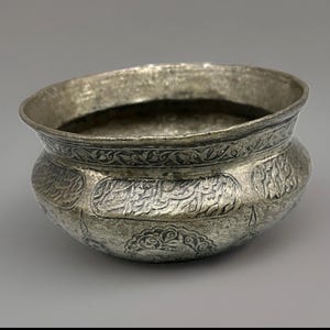 May include: A metallic bowl with a weathered silver finish, featuring an engraved band of intricate floral patterns around the rim and a series of rectangular panels containing embossed calligraphic script and decorative motifs on the body.