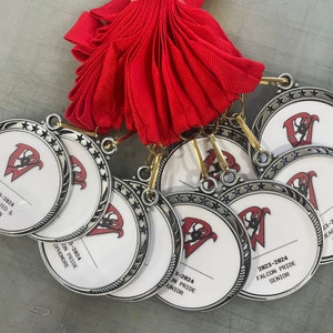 May include: Five silver medals with a black and white logo and text. The medals have a star-shaped border and are stacked on top of each other. The text on the medals reads "2023-2024 Falcon Pride Senior", "2023-2024 Falcon Pride Junior", "2023-2024 Falcon Pride Sophomore", "2023-2024 Dedicated & Driven", and "2023-2024 Heart & Gold".