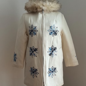 Puede incluir: Abrigo de invierno color crema con cuello de piel sintética y diseños de copos de nieve bordados en tonos azules. El abrigo tiene cierre de cremallera y mangas largas. Una prenda de abrigo elegante.