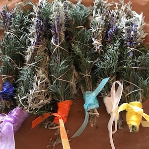 May include: Bundles of dried herbs, including lavender and evergreen sprigs, are tied with twine and colorful ribbons. The ribbons are in shades of blue, purple, orange, white, and yellow. The bundles are arranged on a brown surface.