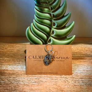 May include: A silver charm with a green and gray stone and a silver heart charm. The charm is attached to a silver chain. The charm is on a brown card with the text "Calm Karma Crystal Healing for Animals" and "Handmade in Cornwall UK".