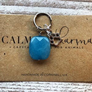 May include: A blue gemstone charm with a silver paw print charm attached to a silver key ring. The charms are on a brown card with the text "CALM karma CRYSTAL HEALING FOR ANIMALS" and "HANDMADE IN CORNWALL U.K."