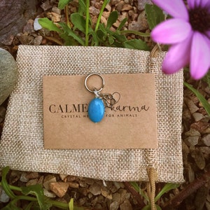 May include: A blue crystal charm with a silver chain and a silver charm that says "karma for animals". The charm is on a brown card that says "Calme Crystal Healing".