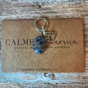 May include: A blue and grey stone charm with a silver paw print charm attached to a silver key ring. The charms are on a brown card with the text "CALME Crystal Karma for Animals" and "HANDMADE IN CORNWALL UK".
