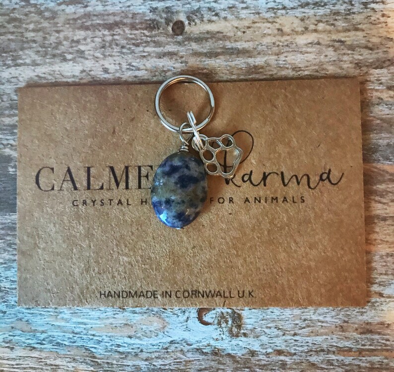 May include: A blue and grey stone charm with a silver paw print charm attached to a silver key ring. The charms are on a brown card with the text "CALME Crystal Karma for Animals" and "HANDMADE IN CORNWALL UK".