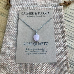 Pode incluir: Colar com pingente de quartzo rosa e corrente prateada, apresentado num cart&atilde;o dentro de um pequeno saco de juta. O cart&atilde;o diz "CALMER & KARMA" e "ROSE QUARTZ". O texto menciona al&iacute;vio do stress e apoio emocional.