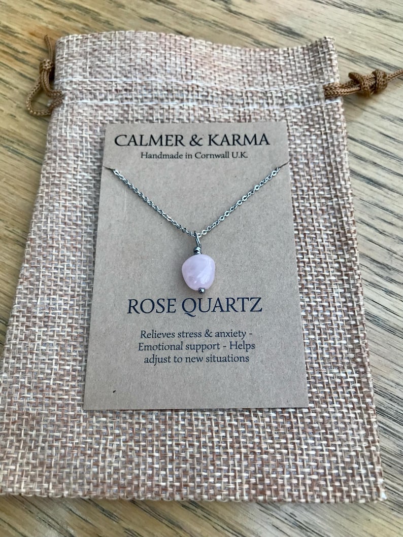 Pode incluir: Colar com pingente de quartzo rosa e corrente prateada, apresentado num cart&atilde;o dentro de um pequeno saco de juta. O cart&atilde;o diz "CALMER & KARMA" e "ROSE QUARTZ". O texto menciona al&iacute;vio do stress e apoio emocional.