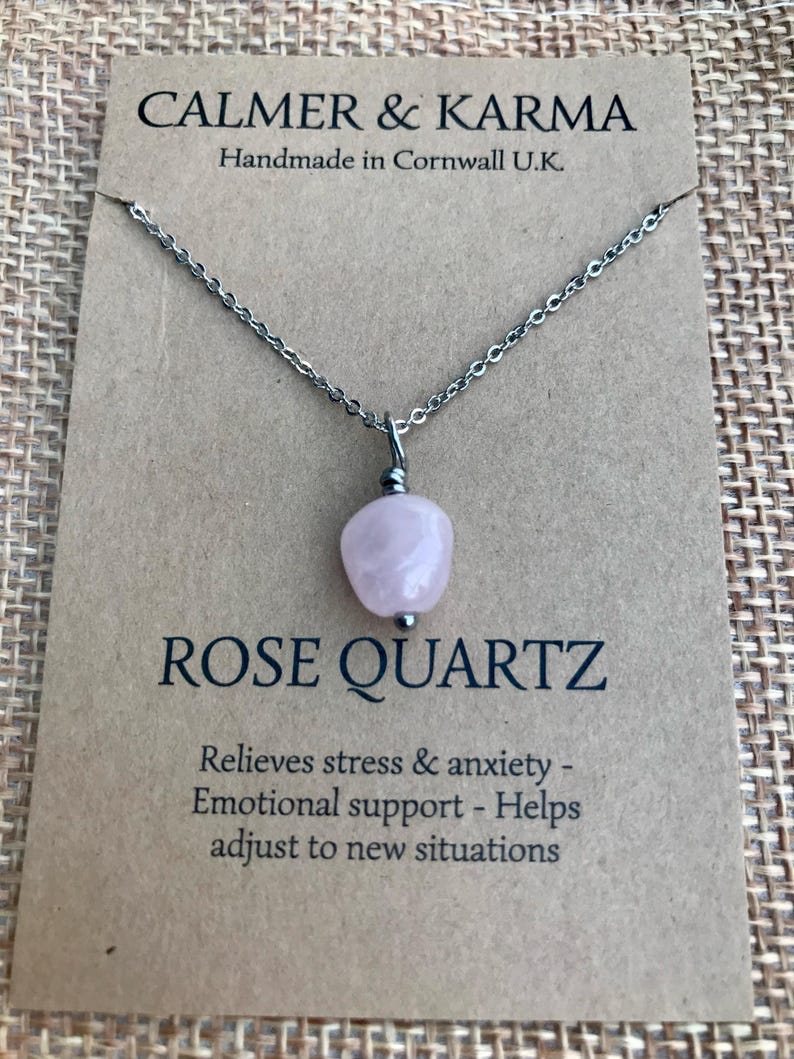 Pode incluir: Um colar com pingente de quartzo rosa e corrente prateada. O pingente &eacute; uma pedra rosa claro polida. O colar &eacute; exibido em um cart&atilde;o marrom com o texto "CALMER & KARMA" e "ROSE QUARTZ". O cart&atilde;o tamb&eacute;m diz "Handmade in Cornwall U.K."