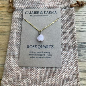 Pode incluir: Um colar com pingente de cora&ccedil;&atilde;o de quartzo rosa em uma corrente dourada, exibido em um cart&atilde;o marrom dentro de uma bolsa bege com cord&atilde;o. O cart&atilde;o diz "CALMER & KARMA" e "ROSE QUARTZ".