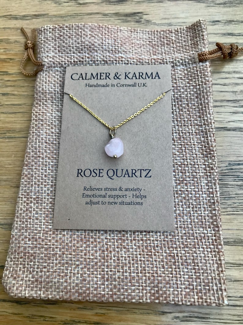 Pode incluir: Um colar com pingente de cora&ccedil;&atilde;o de quartzo rosa em uma corrente dourada, exibido em um cart&atilde;o marrom dentro de uma bolsa bege com cord&atilde;o. O cart&atilde;o diz "CALMER & KARMA" e "ROSE QUARTZ".