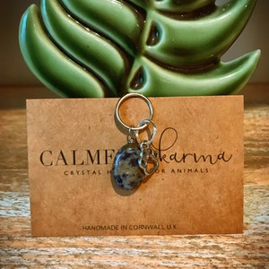 May include: A charm with a grey and black stone and a silver paw print charm. The charm is attached to a gold ring. The charm is on a brown card with the text "CALME karma crystal for animals" and "HANDMADE IN CORNWALL U.K."