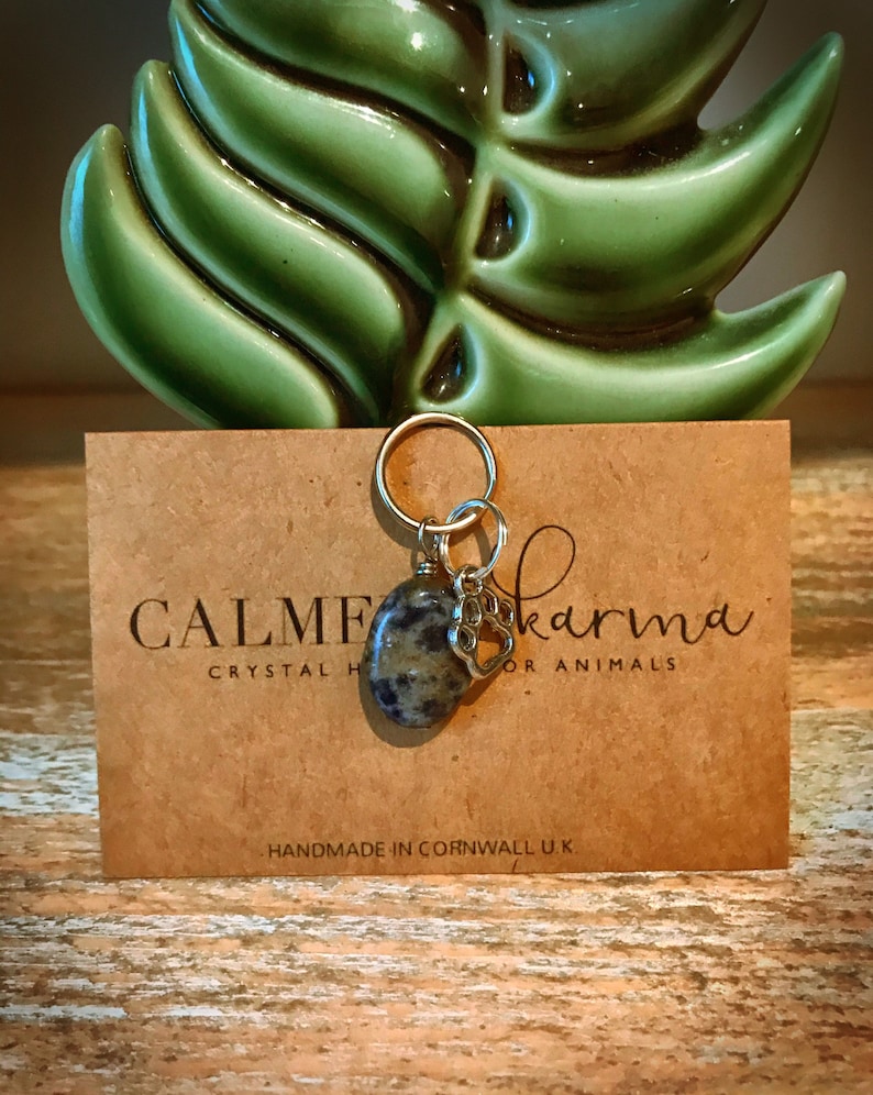May include: A charm with a grey and black stone and a silver paw print charm. The charm is attached to a gold ring. The charm is on a brown card with the text "CALME karma crystal for animals" and "HANDMADE IN CORNWALL U.K."