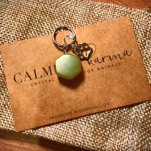 May include: A green crystal charm with a silver paw print charm attached to a silver key ring. The charms are on a brown card with the text "CALM CRYSTAL karma for ANIMALS HANDMADE IN CORNWALL UK".