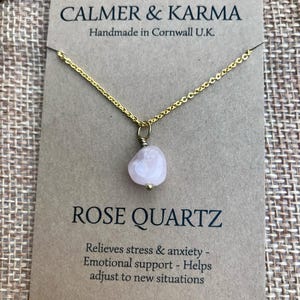 Pode incluir: Um colar com corrente dourada e pingente de quartzo rosa. O pingente &eacute; uma pedra arredondada rosa claro. O colar &eacute; exibido em um cart&atilde;o marrom com o texto "CALMER & KARMA" e "ROSE QUARTZ".