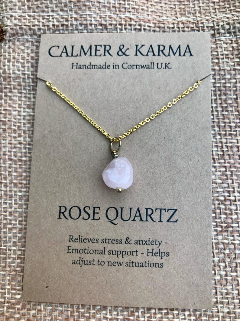 Pode incluir: Um colar com corrente dourada e pingente de quartzo rosa. O pingente &eacute; uma pedra arredondada rosa claro. O colar &eacute; exibido em um cart&atilde;o marrom com o texto "CALMER & KARMA" e "ROSE QUARTZ".