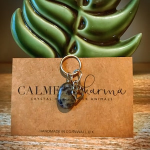 May include: A silver charm with a paw print and a grey and black stone charm on a gold chain. The charm is attached to a brown card with the text "Calm Karma Crystal Healing for Animals" and "Handmade in Cornwall U.K."