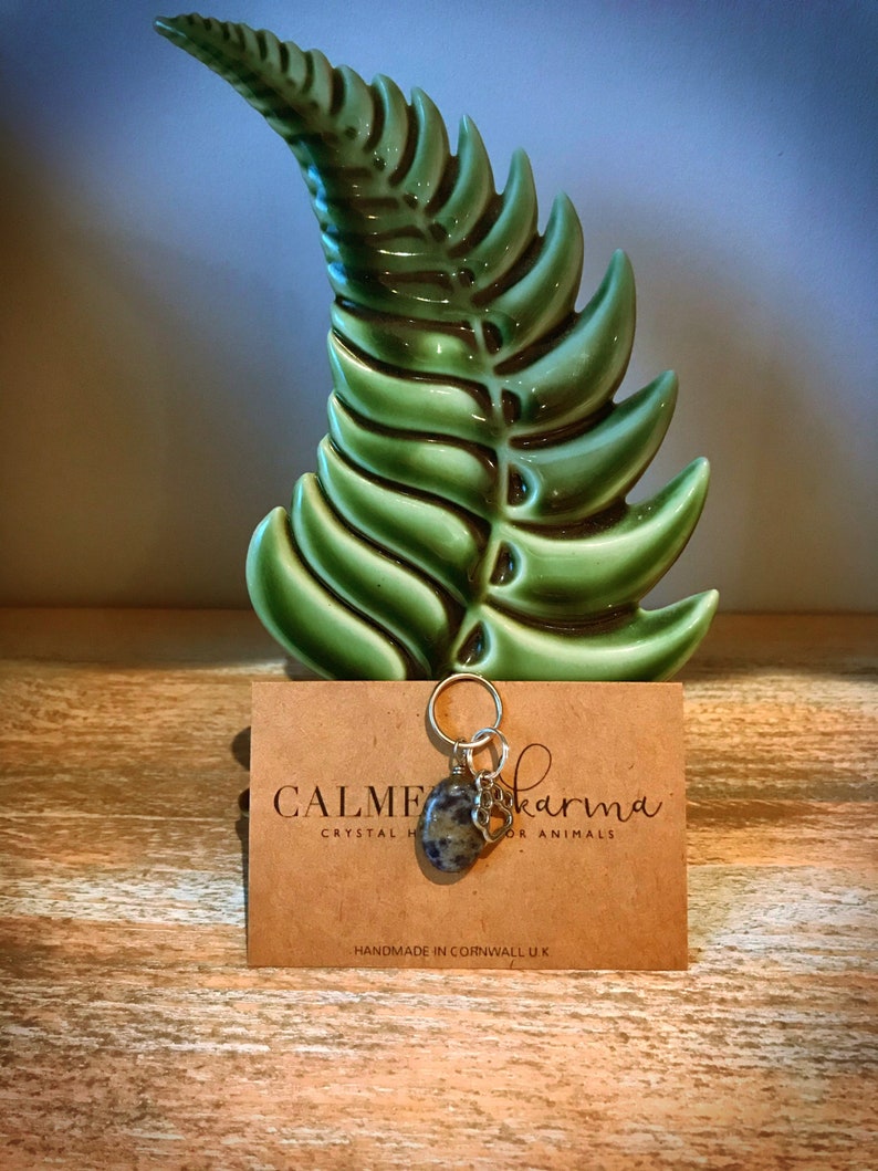 May include: A silver charm with a green and gray stone and a silver heart charm. The charm is attached to a silver chain. The charm is on a brown card with the text "Calm Karma Crystal Healing for Animals" and "Handmade in Cornwall UK".