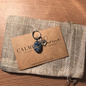 May include: A small blue and white crystal stone charm with a silver paw print charm attached to a silver key ring. The charm is packaged in a small burlap bag with a brown card that reads "Calmer Karma Crystal Healing for Animals" and "Handmade in Cornwall, UK".