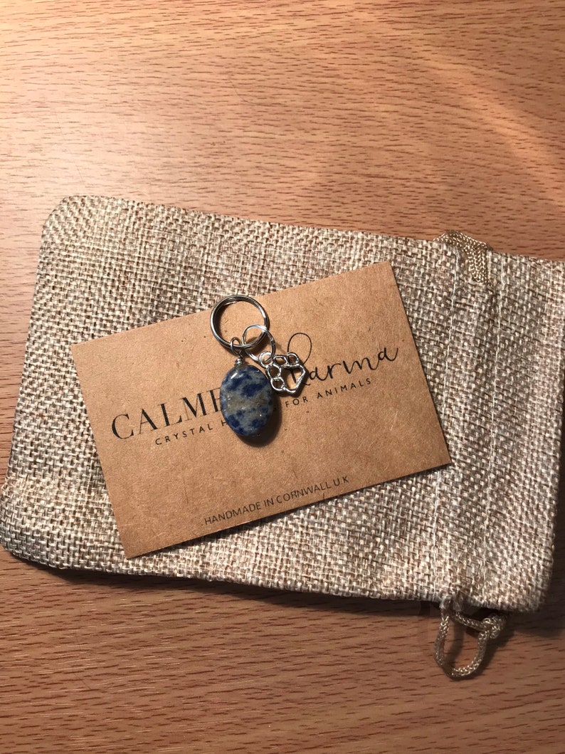 May include: A small blue and white crystal stone charm with a silver paw print charm attached to a silver key ring. The charm is packaged in a small burlap bag with a brown card that reads "Calmer Karma Crystal Healing for Animals" and "Handmade in Cornwall, UK".