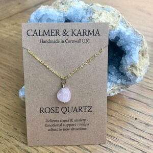 Pode incluir: Um colar de corrente dourada com um pingente de quartzo rosa. O colar &eacute; exibido em um cart&atilde;o marrom com o texto "CALMER & KARMA" e "ROSE QUARTZ". O cart&atilde;o tamb&eacute;m inclui o texto "Handmade in Cornwall U.K."