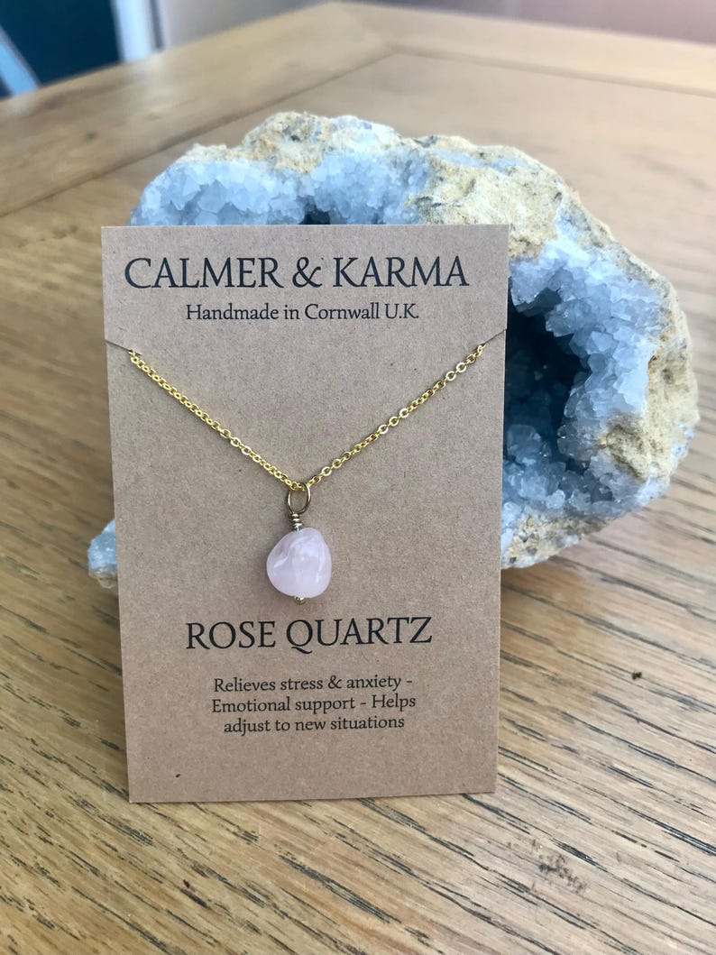 Pode incluir: Um colar de corrente dourada com um pingente de quartzo rosa. O colar &eacute; exibido em um cart&atilde;o marrom com o texto "CALMER & KARMA" e "ROSE QUARTZ". O cart&atilde;o tamb&eacute;m inclui o texto "Handmade in Cornwall U.K."