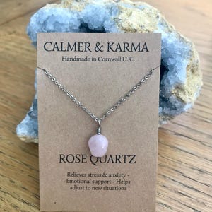 Pode incluir: Um colar com pingente de quartzo rosa em um cart&atilde;o marrom com o texto "CALMER & KARMA" e "ROSE QUARTZ". O cart&atilde;o tamb&eacute;m diz "Handmade in Cornwall U.K." e lista os benef&iacute;cios do quartzo rosa. O colar &eacute; exibido em frente a um cristal azul.