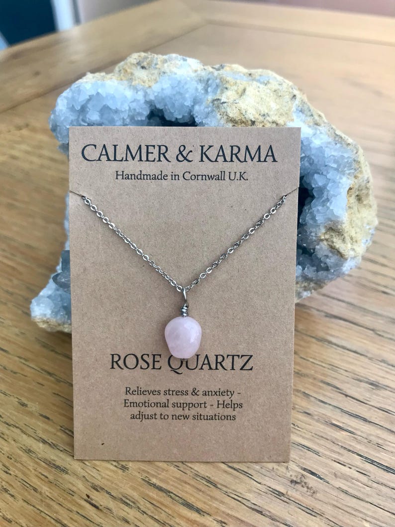 Pode incluir: Um colar com pingente de quartzo rosa em um cart&atilde;o marrom com o texto "CALMER & KARMA" e "ROSE QUARTZ". O cart&atilde;o tamb&eacute;m diz "Handmade in Cornwall U.K." e lista os benef&iacute;cios do quartzo rosa. O colar &eacute; exibido em frente a um cristal azul.