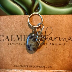 May include: A silver charm with a paw print hangs from a gold key ring. A round, gray and white stone is attached to the ring. The charm and stone are on a brown card with the text "CALME Karma Crystal Healing for Animals" and "Handmade in Cornwall U.K."