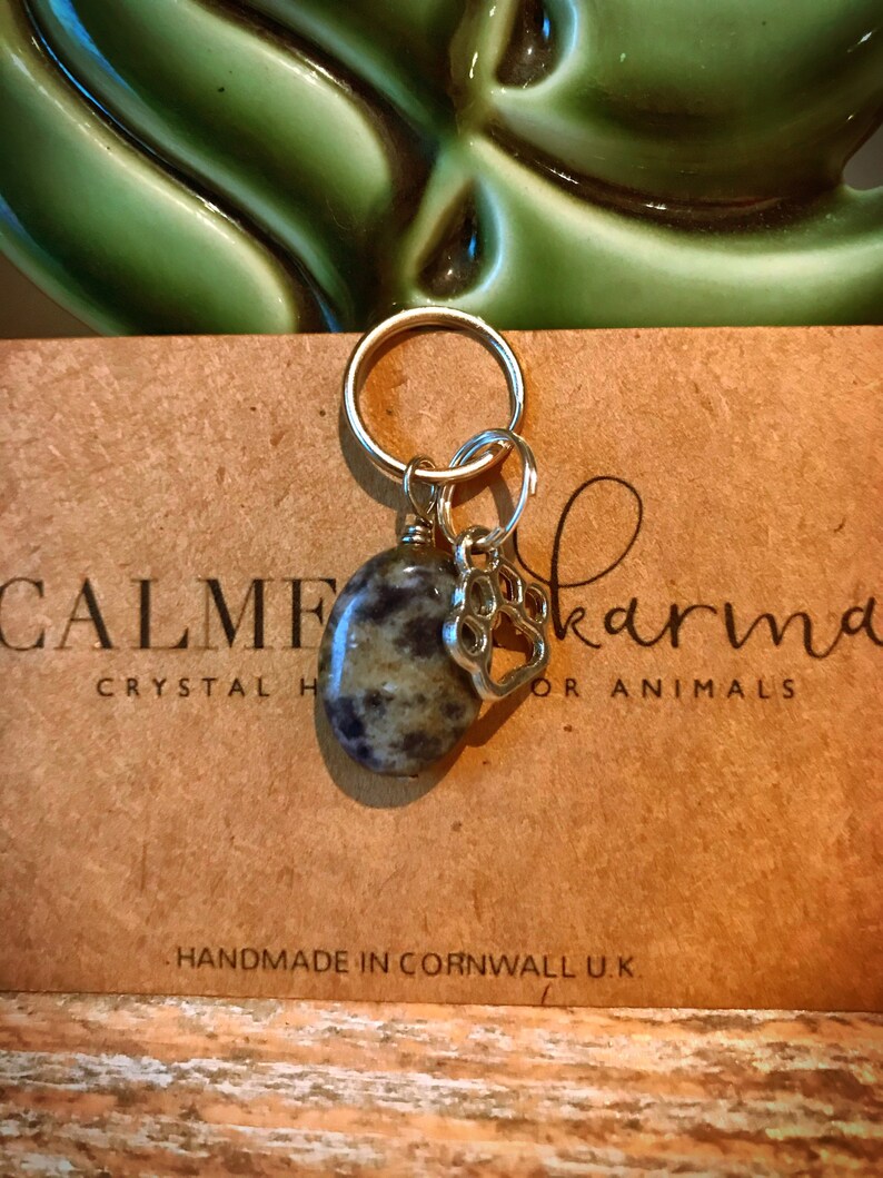 May include: A silver charm with a paw print hangs from a gold key ring. A round, gray and white stone is attached to the ring. The charm and stone are on a brown card with the text "CALME Karma Crystal Healing for Animals" and "Handmade in Cornwall U.K."