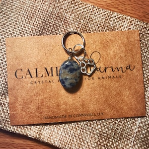 May include: A blue and white stone charm with a silver paw print charm attached to a silver key ring. The charms are on a brown card with the text "Calm Crystal Karma for Animals" and "Handmade in Cornwall UK".