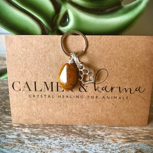 May include: A brown tiger's eye crystal charm with a silver paw print charm attached to a silver key ring. The charms are on a brown card with the text "Calmer & Karma Crystal Healing for Animals".