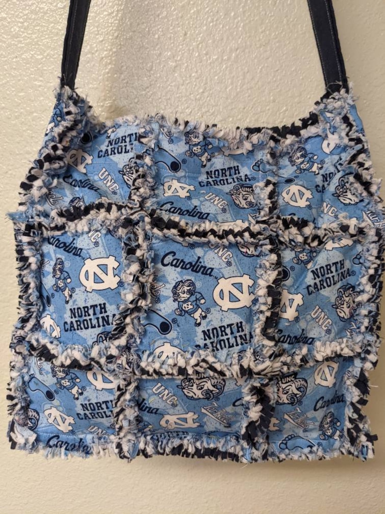 May include: A blue and white patchwork tote bag with a frayed edge. The bag features a repeated pattern of the University of North Carolina Tar Heels logo, mascot, and the words "North Carolina" and "UNC".