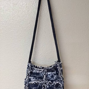 May include: A black and white fabric crossbody bag with a frayed edge. The bag features a patchwork design with the Dallas Cowboys logo and the words "Cowboys" and "Boys" repeated throughout.