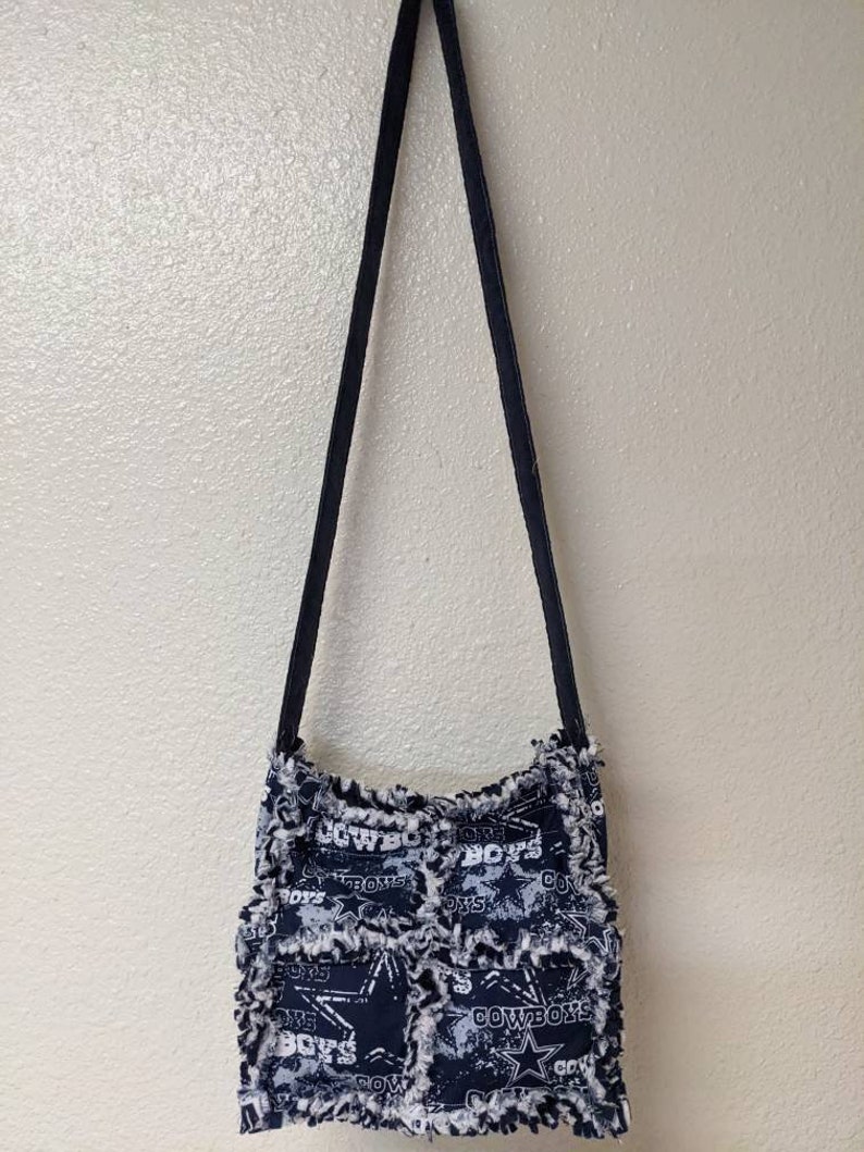 May include: A black and white fabric crossbody bag with a frayed edge. The bag features a patchwork design with the Dallas Cowboys logo and the words "Cowboys" and "Boys" repeated throughout.