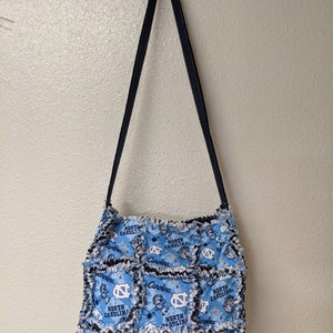 May include: A blue and white patchwork tote bag with a black strap. The bag is made of fabric squares with the North Carolina Tar Heels logo and the words "North Carolina" repeated on each square.