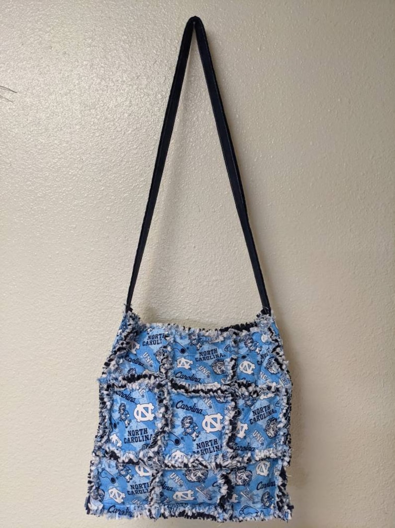 May include: A blue and white patchwork tote bag with a black strap. The bag is made of fabric squares with the North Carolina Tar Heels logo and the words "North Carolina" repeated on each square.