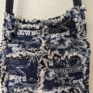 May include: A blue and white patchwork fabric crossbody bag with a frayed edge. The bag features a repeated pattern of the Dallas Cowboys logo and the word "Cowboys".
