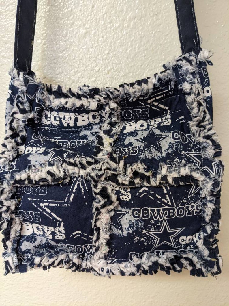 May include: A blue and white patchwork fabric crossbody bag with a frayed edge. The bag features a repeated pattern of the Dallas Cowboys logo and the word "Cowboys".