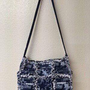 May include: A blue and white patchwork purse with a frayed edge. The purse has a long strap and features the Dallas Cowboys logo and the words "Cowboys" and "Wboys".