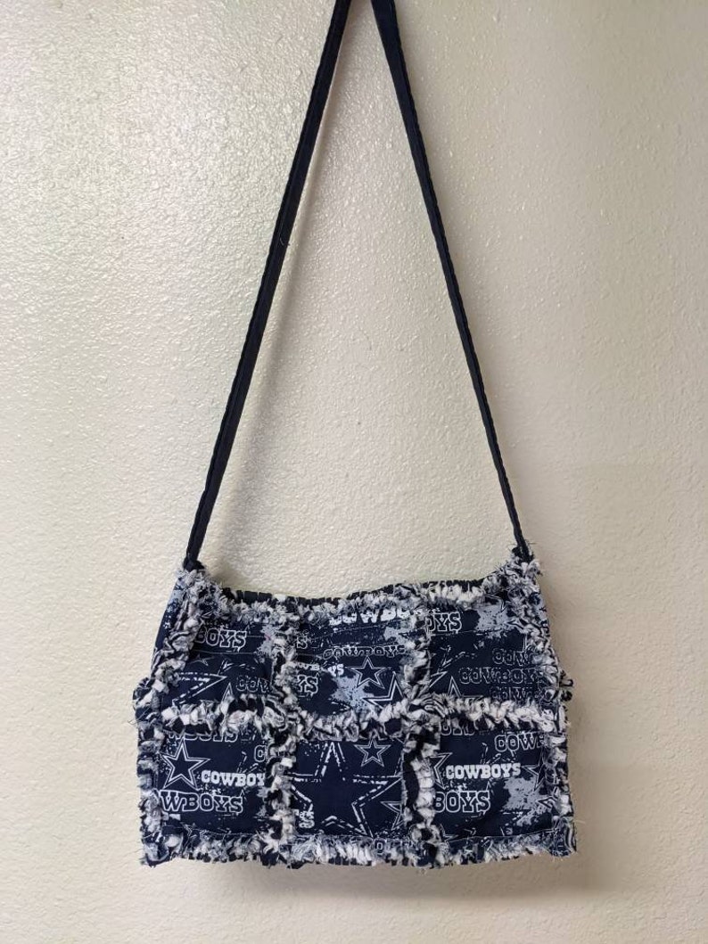 May include: A blue and white patchwork purse with a frayed edge. The purse has a long strap and features the Dallas Cowboys logo and the words "Cowboys" and "Wboys".
