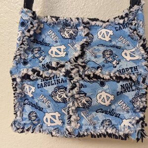 May include: A blue and white fabric tote bag with a frayed edge. The bag features a repeated pattern of the UNC Tar Heels logo, the words "UNC" and "Carolina", and a ram mascot. The bag has a black strap.