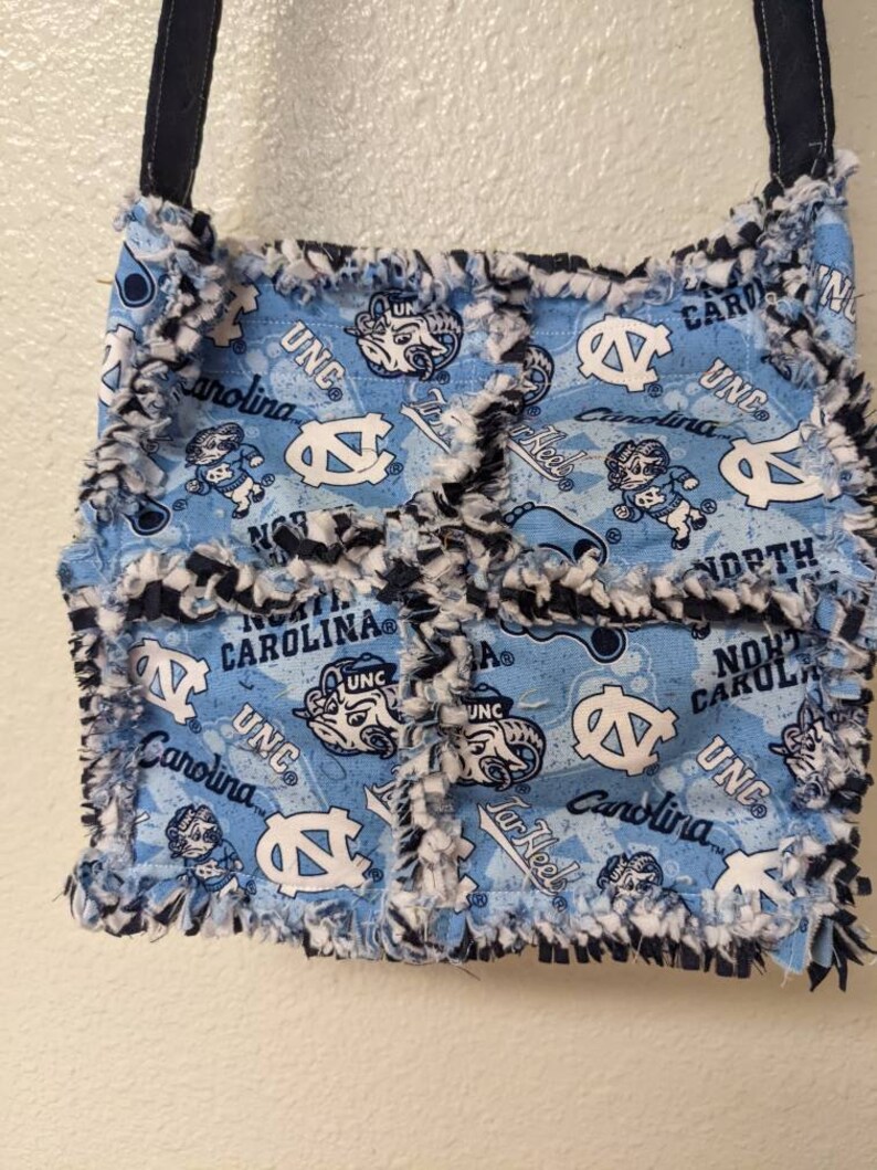 May include: A blue and white fabric tote bag with a frayed edge. The bag features a repeated pattern of the UNC Tar Heels logo, the words "UNC" and "Carolina", and a ram mascot. The bag has a black strap.