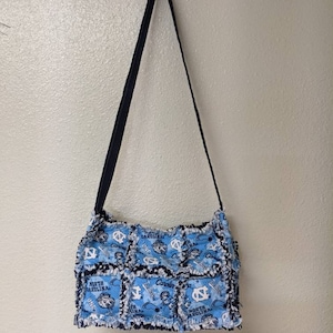 May include: A blue and white patchwork tote bag with a black strap. The bag features a repeated pattern of the University of North Carolina Tar Heels logo and the words "North Carolina".